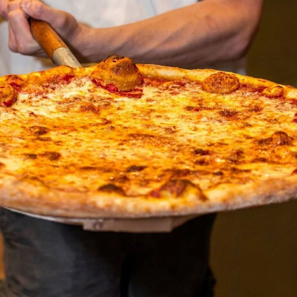A chef holds a large, freshly baked cheese pizza on a wooden peel, ready to serve.