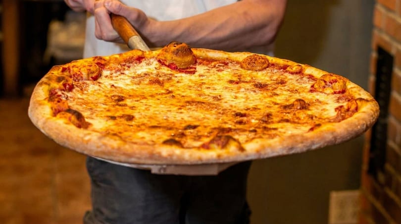 A chef holds a large, freshly baked cheese pizza on a wooden peel, ready to serve.